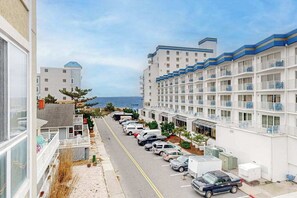 A coastal hotel with multiple-story buildings, a parking lot with cars, and a view of the ocean. - Family-Friendly Beach Condo: Balcony, w/d, shared outdoor pool & gym (Ocean City)