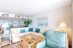 A living room with a sofa, coffee table, and a chair. There is a plant, a painting on the wall, and a lamp. - Irene 607 (Ocean City)