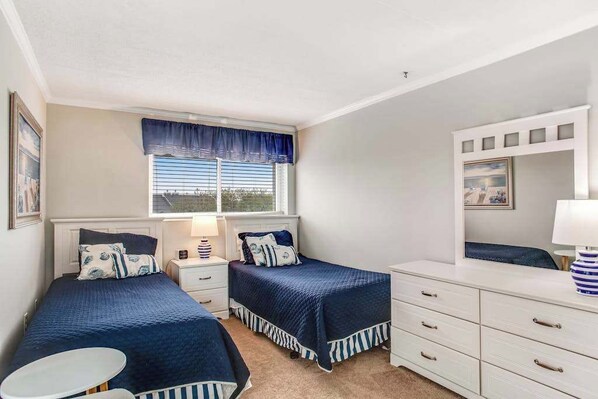 A bedroom with two beds, a window with blinds, a bedside table with a lamp, a dresser, and a mirror. - Convenient & comfortable condo with pool & water views from balcony (Ocean City)