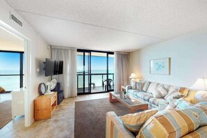 A living room with a sofa set, a flat-screen TV, a coffee table, and a view of the sea through large windows. - Darling oceanfront condo with balcony, W/D, AC, pools & game room (Ocean City)
