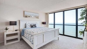 Une chambre avec une grande fenêtre, un lit, une table de chevet et une commode.