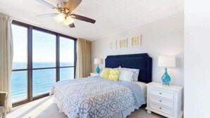 A bedroom with a bed, nightstands, a ceiling fan, and a large window overlooking the ocean. - Updated oceanfront condo with balcony, AC, W/D, pools & sauna (Ocean City)