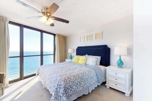 Ein Schlafzimmer mit Bett, Nachttischen, Deckenventilator und großem Fenster mit Blick auf das Meer.