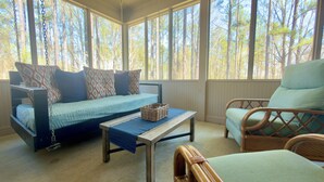 A room with a bed, a chair, a table, and large windows with a view of trees.