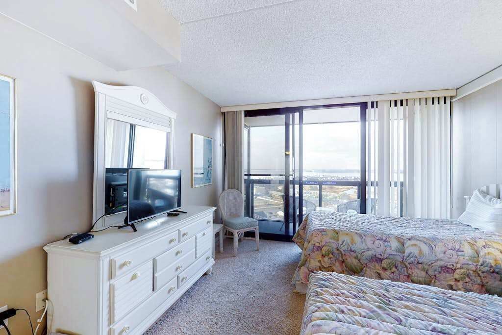 Ein Hotelzimmer mit einem großen Bett, einem Fernseher auf einem Schminktisch, einem Sessel und einem Balkon mit Aussicht.
