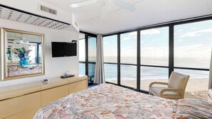 Een slaapkamer met een groot bed, een televisie, een stoel en uitzicht op het strand.