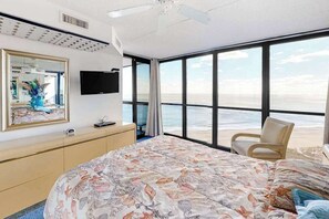 A bedroom with a large bed, a TV, a chair, and a view of the beach. - Bright & convenient condo with pool, balcony & angled ocean views (Ocean City)
