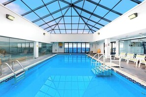 Une piscine intérieure dotée d’un toit en verre, offrant une vue dégagée sur la mer, et des chaises longues disposées le long du bord de la piscine.