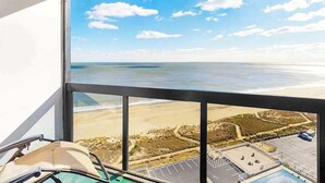 Een kamer met een groot raam dat uitkijkt op een strand en de oceaan, een zwembad en een parkeerplaats.