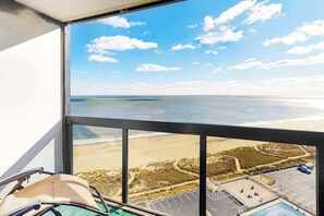 A room with a large window overlooking a beach and ocean, a pool, and a parking area. - Bright & convenient condo with pool, balcony & angled ocean views (Ocean City)