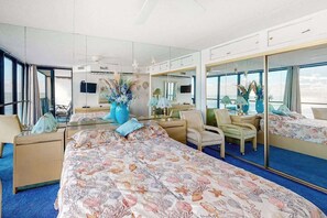 Une chambre d’hôtel avec un grand lit, un coin salon comprenant un fauteuil et un bureau, un grand miroir et une vue sur la mer.