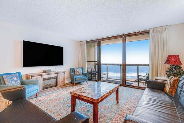 A living room with a large TV, a sofa, a coffee table, and a view of the ocean. - Convenient beachfront condo with angled views, cozy balcony, pools, gym & tennis (Ocean City)
