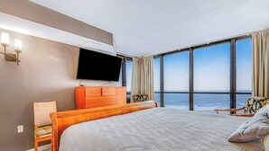 A hotel room with a bed, a TV, a dresser, and a view of the ocean. - Convenient beachfront condo with angled views, cozy balcony, pools, gym & tennis (Ocean City)