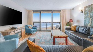 A living room with a large TV, a sofa, a coffee table, and a view of the ocean. - Convenient beachfront condo with angled views, cozy balcony, pools, gym & tennis (Ocean City)