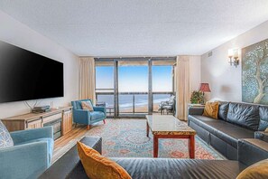 A living room with a large TV, a sofa, a coffee table, and a view of the ocean. - Convenient beachfront condo with angled views, cozy balcony, pools, gym & tennis (Ocean City)