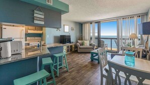 A modern kitchen with a bar area, a dining table with chairs, a sofa, and a view of the ocean. - Updated condo with balcony, pools, gym, tennis court & AC (Ocean City)