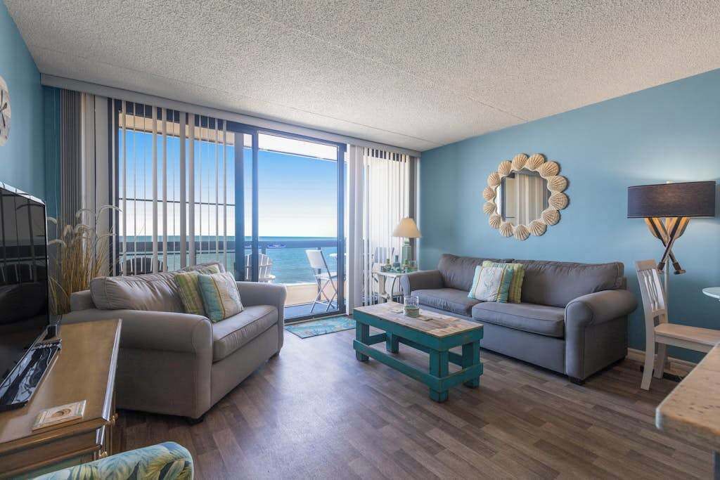 Uma sala de estar com dois sofás, uma mesa de centro, uma televisão e uma janela grande com vista para o mar.