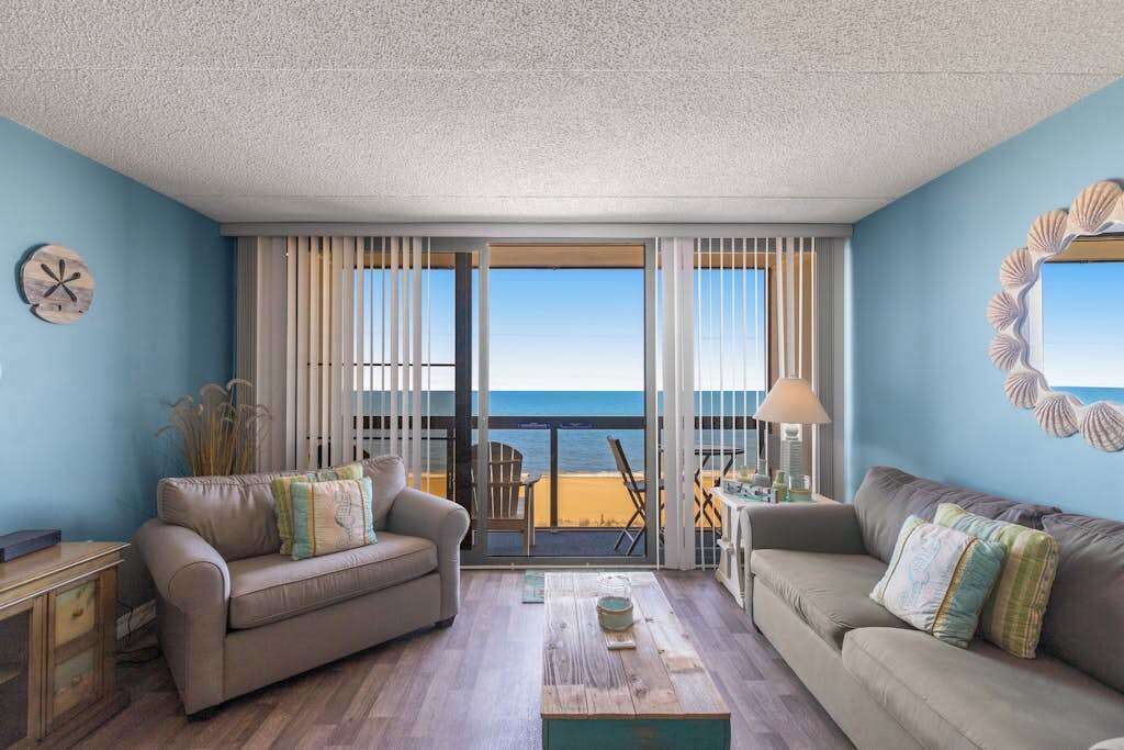 A room with two sofas, a coffee table, and a view of the beach through large windows.