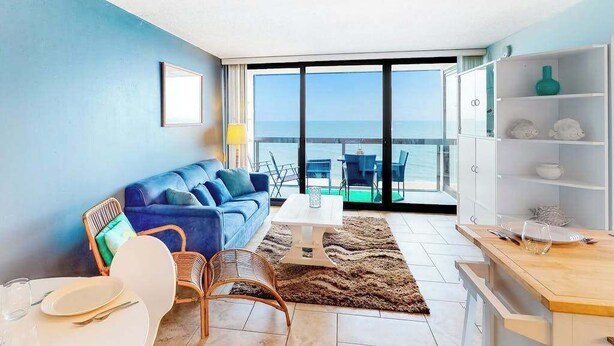 A modern living room with a blue sofa, a dining table, and a view of the sea through large windows. - Delightful, well-equipped beachfront condo with balcony, views, pools & gym (Ocean City)