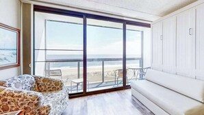 A spacious room with a large sliding glass door leading to a balcony, a white sofa, and a patterned armchair. - Budget-friendly beachfront studio with balcony, tennis, gym, pools & sauna (Ocean City)