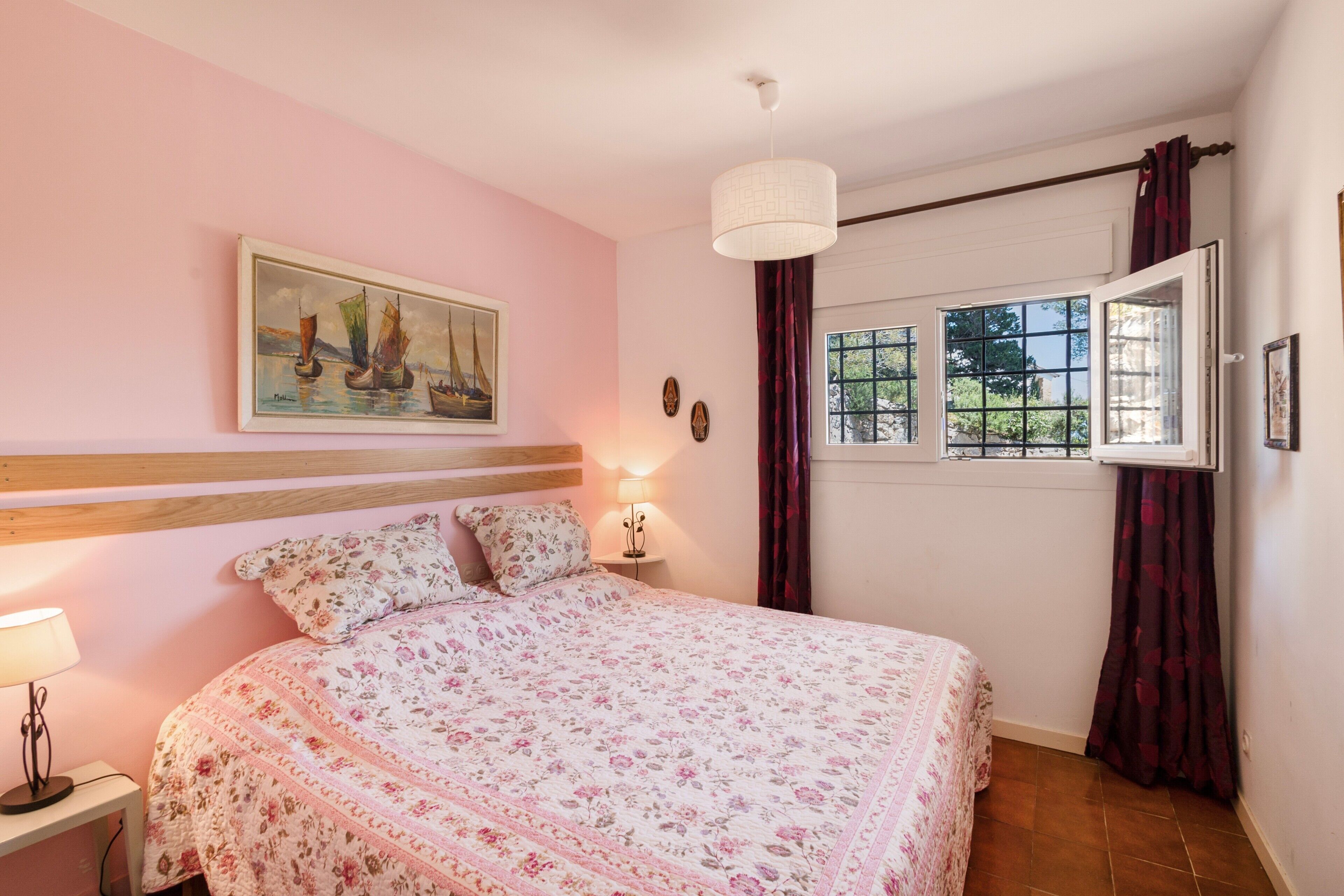 A bedroom with a bed, a nightstand, a lamp, a painting, a window with curtains, and a view of greenery outside.