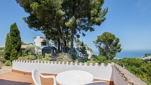 Eine Terrasse mit Tisch und Stühlen mit Blick auf eine Küstenlandschaft mit Bäumen und Gebäuden.