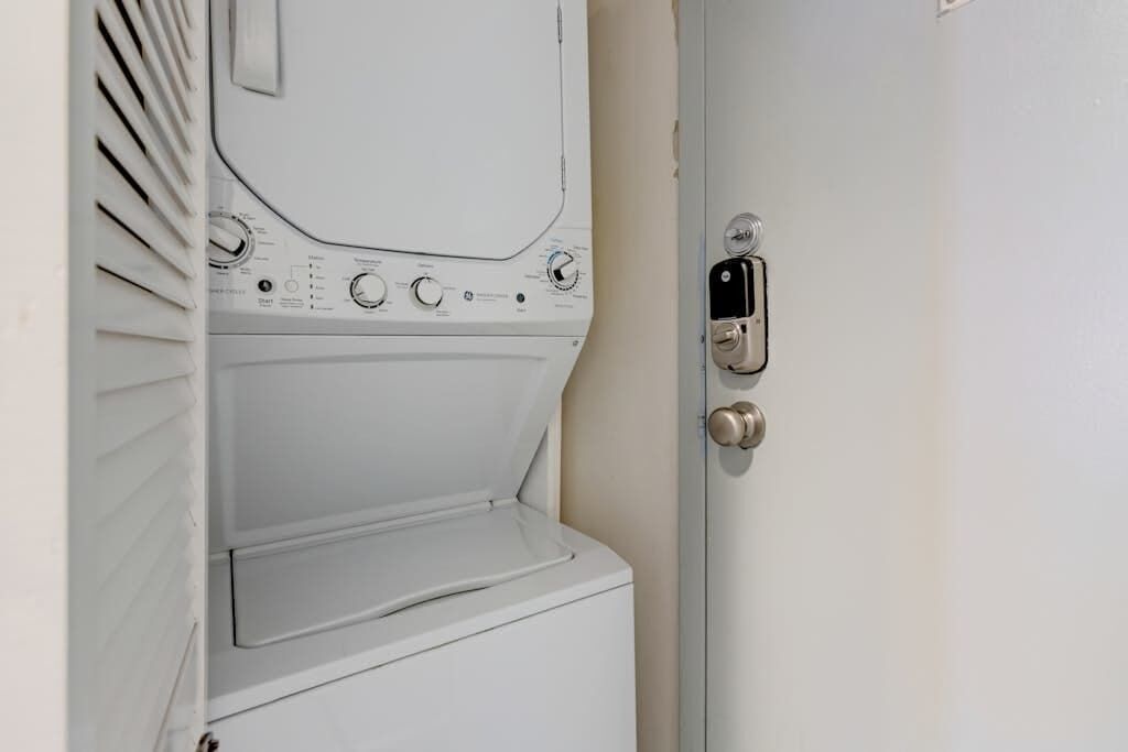 Lavadora blanca con perillas de control y pantalla digital, fijada a la pared junto a una puerta con cerradura.