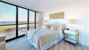 A bedroom with a large window, a bed, a bedside table, and a wall-mounted artwork. - Cozy beachfront condo with balcony, AC, pools, game room & gym (Ocean City)