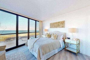 A bedroom with a large window, a bed, a bedside table, and a wall-mounted artwork. - Cozy beachfront condo with balcony, AC, pools, game room & gym (Ocean City)