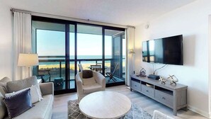 A modern hotel room with a balcony, a flat-screen TV, a sofa, a chair, and a small table. - Cozy beachfront condo with balcony, AC, pools, game room & gym (Ocean City)