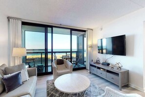 A modern hotel room with a balcony, a flat-screen TV, a sofa, a chair, and a small table. - Cozy beachfront condo with balcony, AC, pools, game room & gym (Ocean City)
