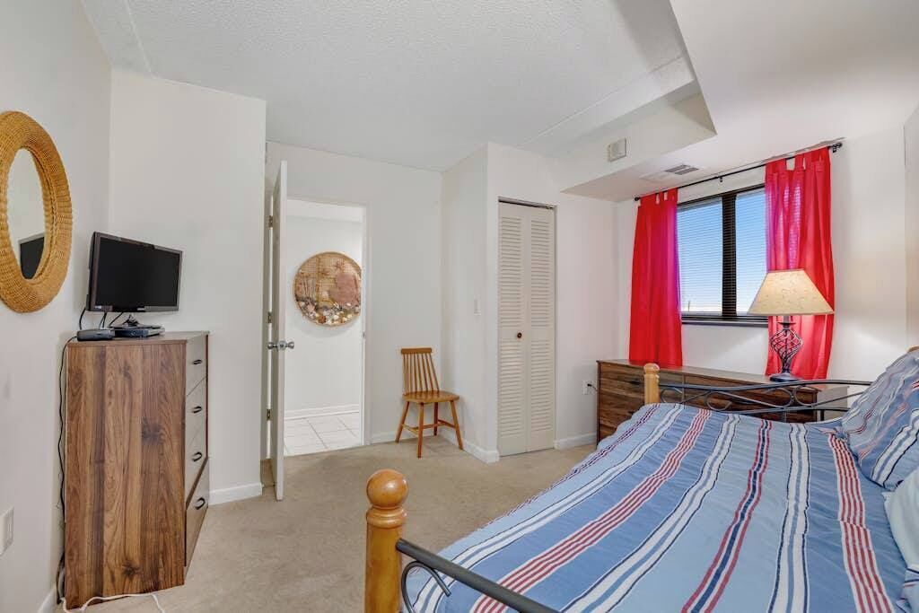 A bedroom with a bed, a TV on a wooden cabinet, a chair, a wardrobe, and a window with red curtains.