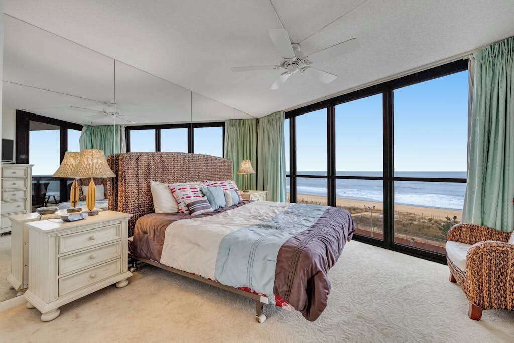 A bedroom with a large bed, a dresser, a nightstand, and a view of the beach.