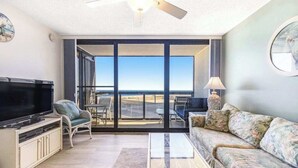 A living room with a television, sofa, and chairs, a ceiling fan, and a large sliding glass door leading to a balcony.