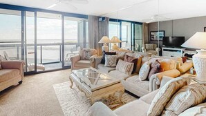 A spacious living room with a large window, a ceiling fan, and a television. - Oceanfront condo with balcony, washer/dryer, pools, & gym (Ocean City)
