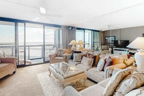 A spacious living room with a large window, a ceiling fan, and a television. - Oceanfront condo with balcony, washer/dryer, pools, & gym (Ocean City)