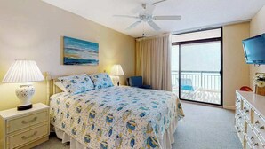 Hotelkamer met een bed, twee bedlampen, een plafondventilator, een televisie, een balkon met een stoel en een schilderij aan de muur.