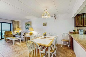 Ein modernes Wohnzimmer mit Esstisch, weißen Stühlen, einer Couch und einer Küchenzeile inklusive Schränke und Kaffeemaschine.