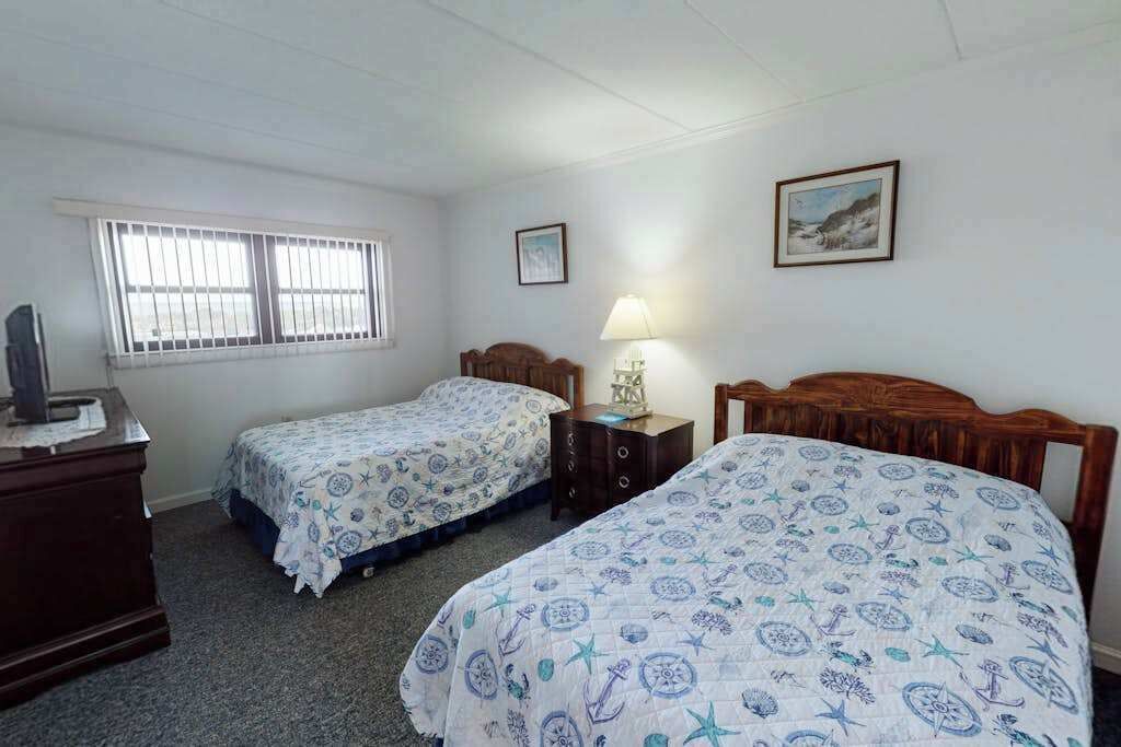 Une chambre avec deux lits, une commode, une lampe et une fenêtre avec des barreaux.
