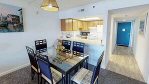 A dining area with a glass table and chairs, a kitchen in the background, and a hallway leading to a blue door. - Beachfront Suite w/Ocean Views From Balcony, Free WiFi, Central AC, Private W/D (Ocean City)