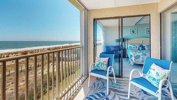 Een balkon met uitzicht op het strand, een bed met bloemenbeddengoed en een blauw-wit gestreept tapijt.
