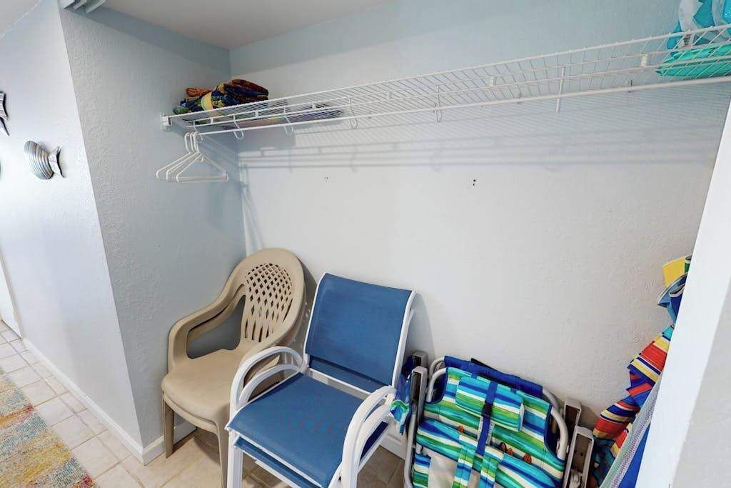 A corner room with a chair, a blue cushioned seat, a white shelf with hanging clothes, and a patterned rug.