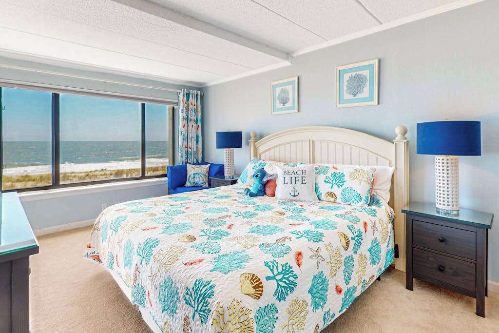 A bedroom with a bed, two nightstands, a window with a view of the beach, and two wall art pieces.