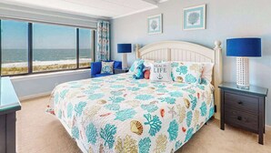 Een slaapkamer met een bed, twee nachtkastjes, een raam met uitzicht op het strand en twee decoratieve wandpanelen.