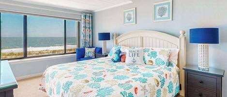A bedroom with a bed, two nightstands, a window with a view of the beach, and two wall art pieces.
