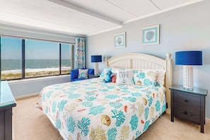 Ein Schlafzimmer mit einem Bett, zwei Nachttischen, einem Fenster mit Blick auf den Strand und zwei Wandbildern.