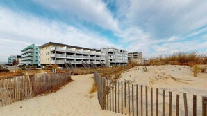 Een hotel aan het strand met meerdere verdiepingen, een zandpad en een houten omheining.