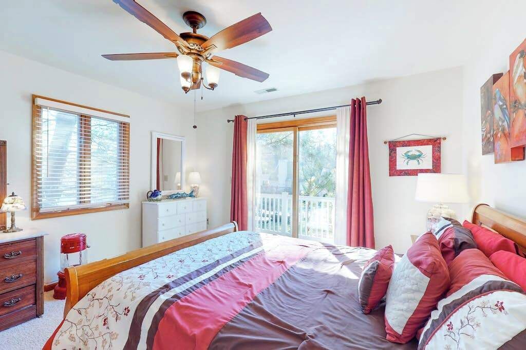 A bedroom with a bed, a ceiling fan, a dresser, a window with blinds, a door, and a balcony with a view.