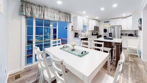 A modern kitchen with white cabinetry, a dining area with a white table and chairs, and a large window with blue shutters. - Endless Summer (Ocean City)