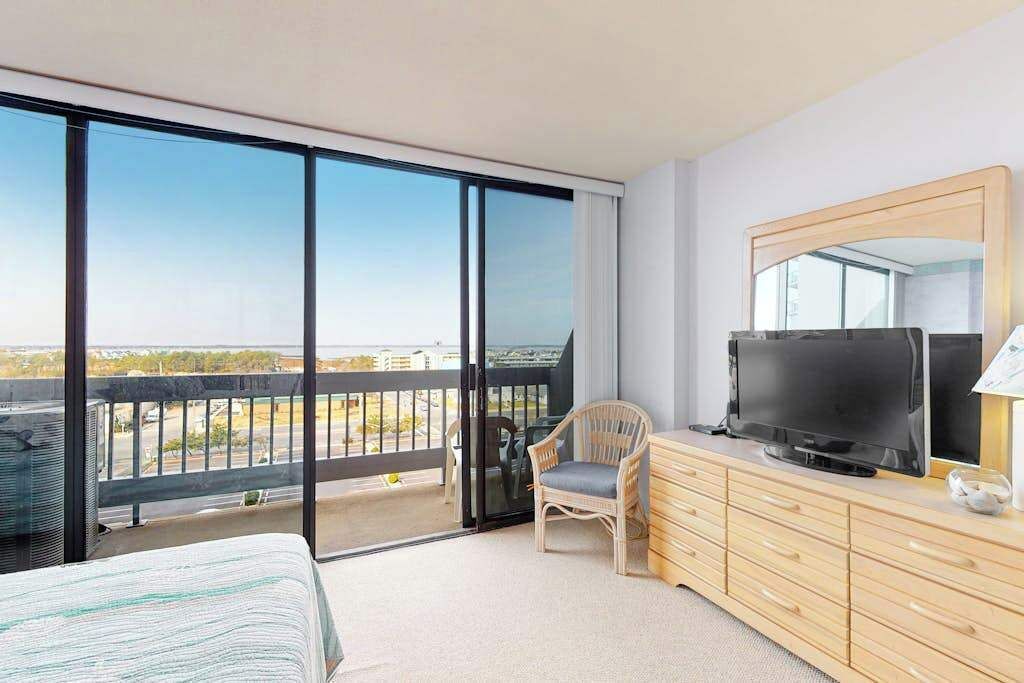 Habitación de hotel con un amplio ventanal corredero que da a un balcón, un televisor sobre una cómoda de madera y una sola silla.